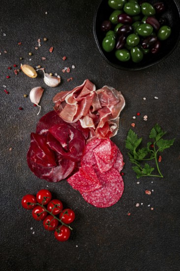 Assorted dried meat, fresh tomatoes and olives spread out on a dark surface, food composition, top