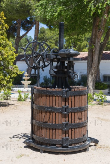 Historische Weinpresse aus Holz und Metall in einem sonnigen Garten mit Bäumen und einem Brunnen,