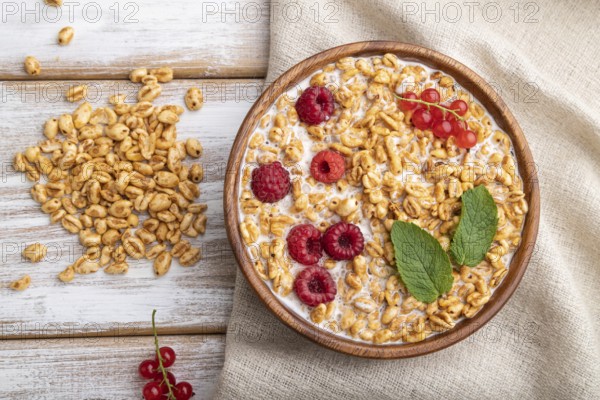 Wheat flakes porridge with milk, raspberry and currant in wooden bowl on white wooden background
