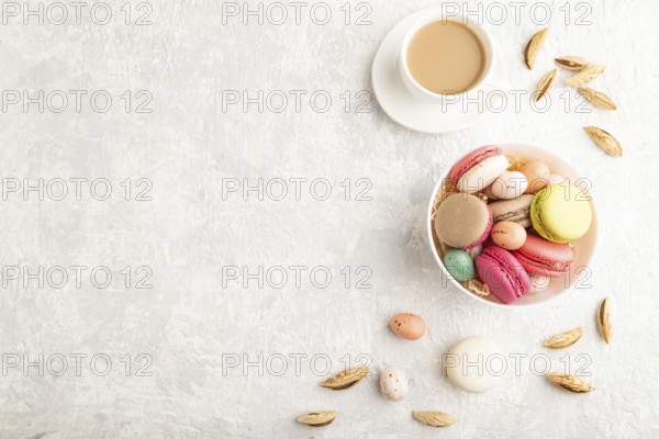 Multicolored macaroons and chocolate eggs in ceramic bowl, cup of coffee on gray concrete
