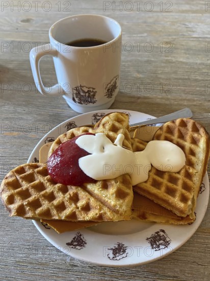 Waffles with jam, cream and cheese on a plate, next to a cup of coffee, served on a wooden table,