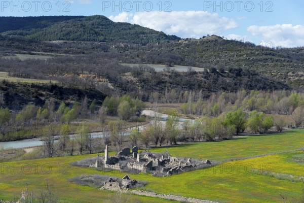 Ruins of an old building in a green field surrounded by a river and wooded hills on a clear spring