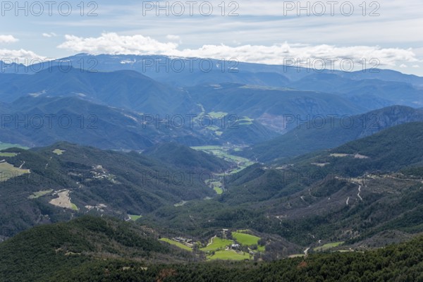 Weitreichende Berglandschaft mit einem grünen Tal, bewaldeten Hängen und einem blauen Himmel mit