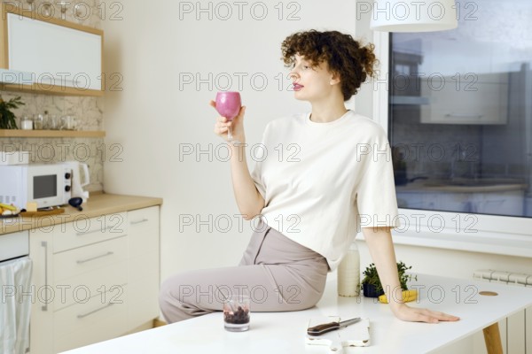 Dreamy and satisfied young woman sits on a kitchen table and holds a glass of smoothie in hand