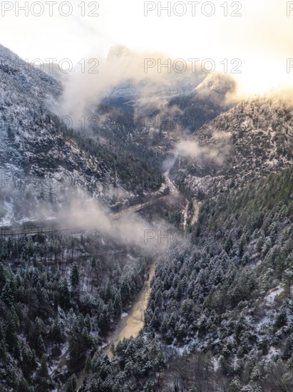 A valley with a river flowing through it surrounded by snow-covered mountains and wafts of mist,