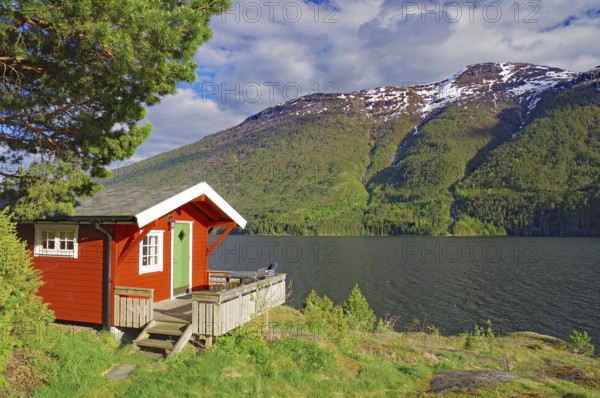 Red-painted cabin on the shore of a fjord against a backdrop of mountains and forest, holiday