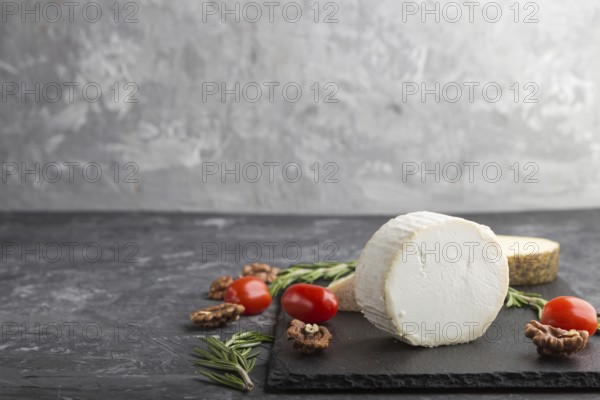 White goat cheese and various types of cheese with rosemary and tomatoes on black slate board on a