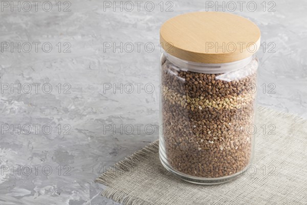 Glass jar with buckwheat poured in layers on a gray concrete background. side view, close up, copy