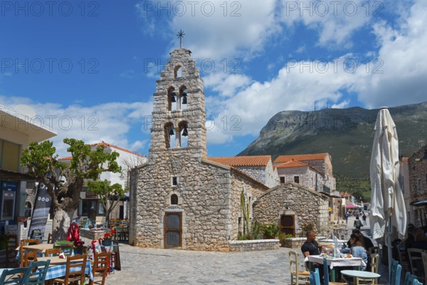 Small church with bell tower in a lively street with cafés and mountains in the background, double