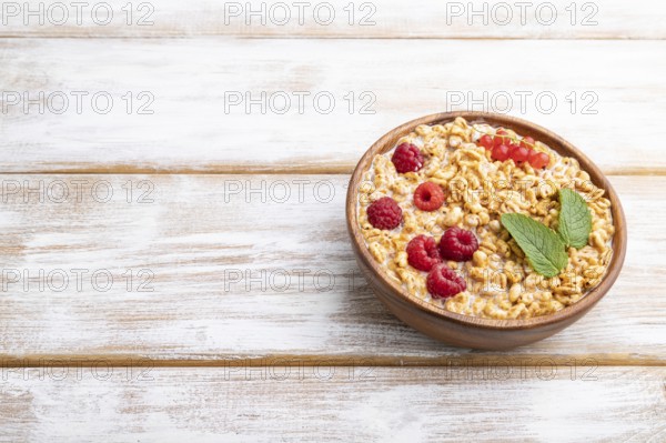 Wheat flakes porridge with milk, raspberry and currant in wooden bowl on white wooden background.