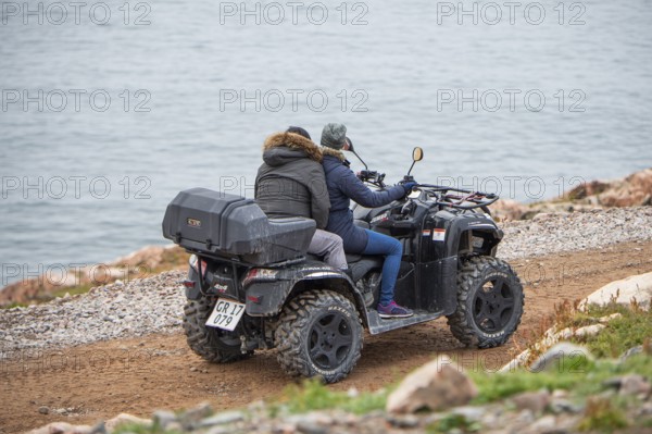 Two people ride a quad bike along a narrow gravel track near the coast, fjord, remote Arctic Inuit