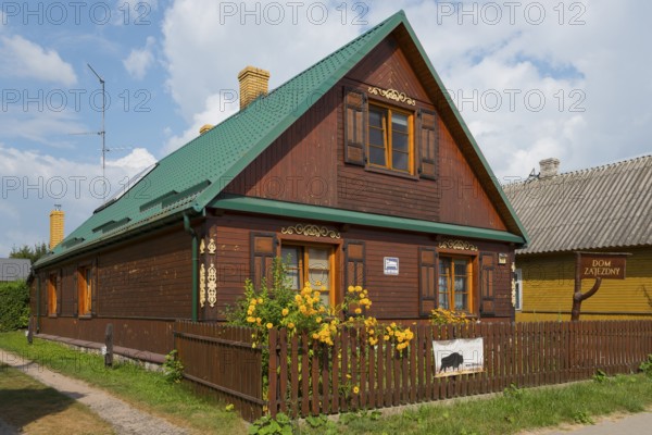 Dark brown wooden house with green roof and flowers in the garden behind the fence, Bialowieza,