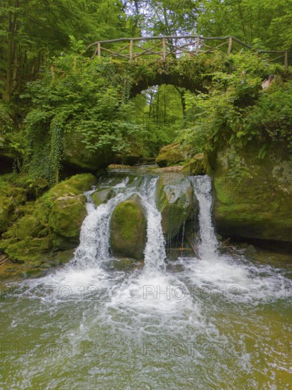 An idyllic waterfall flows under a rustic bridge, surrounded by lush greenery and rocks, aerial
