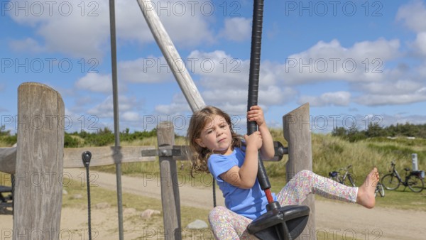 A child happily jumps on a zip wire in a wide, sunny playground setting with a cloudy, blue sky