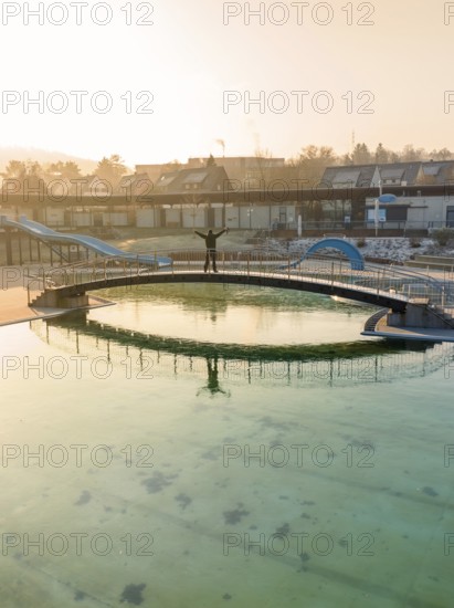 A person stands on a bridge over a swimming pool with green frozen water in the winter sun, ENCW