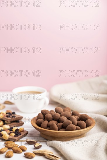 Almond in chocolate dragees in wooden plate and a cup of coffee on white and pink background and
