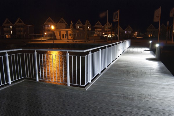 Pier with illuminated lamps, railings and light reflections in the water at night, flags and houses