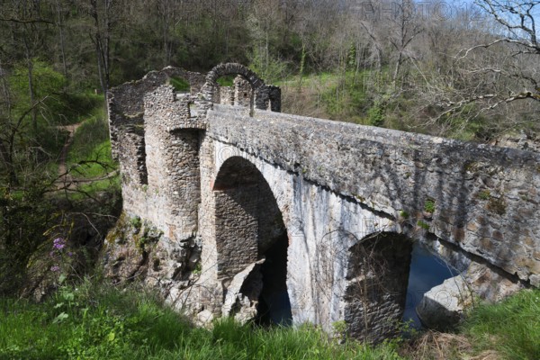 Alte Steinbrücke über einen Fluss, umgeben von bewaldeter Landschaft und Ruinenresten im Frühling,