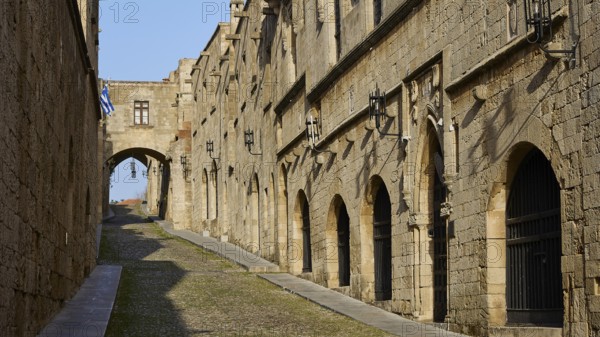 A historic alleyway with stone buildings and arches, blue and white flags in the background,