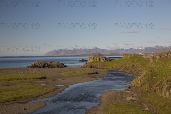 A creek flowing through green meadows at the coast with the ocean and blue sky in the background.