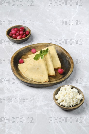 Three golden crepes are elegantly arranged on a wooden plate, accompanied by small bowls of fresh
