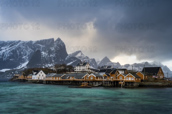 Traditional yellow rorbuer cabins on Sakrisøy Island, snow-capped rocky mountains in the back,