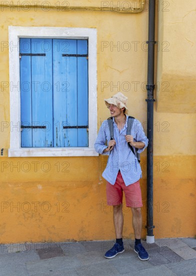 Young man with backpack in front of yellow house wall, yellow house facade with windows with blue