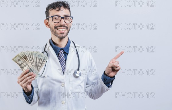 Happy young doctor holding money and pointing to side. Handsome smiling doctor showing money and