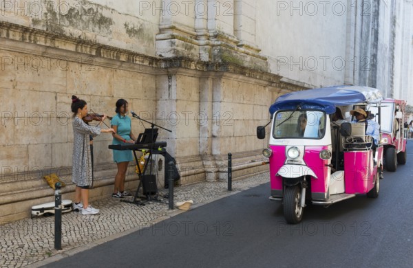 A violinist and a singer play music in front of an old wall while a pink tuk-tuk drives by, Old