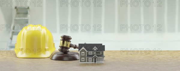 Model house, gavel and yellow hard hat resting on a wood surface in front of drywall and ladder