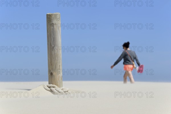 Boundary post on the beach, rope blown over by sand, person crossing the sand drifts, blue sky,