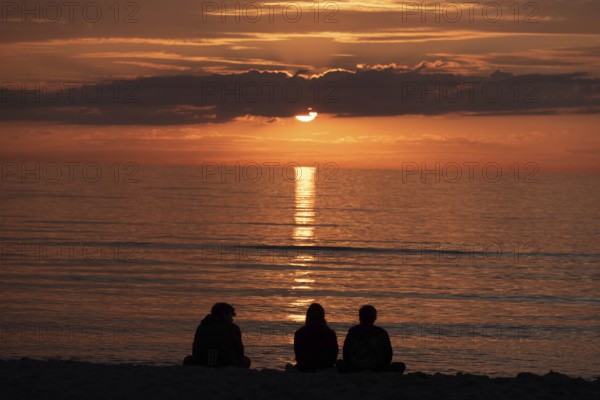 Three people sitting on the beach watching the sunset over the sea, silhouettes in front of warm