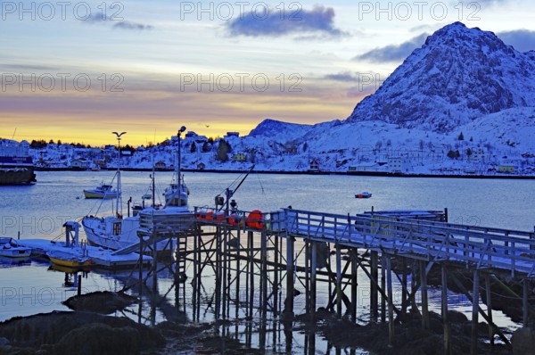 Sunset over a snowy harbour with boats, calm water and snow-capped mountains in the background,
