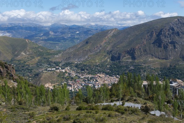 View over a valley with a village surrounded by mountains, lots of greenery and a cloudy sky,