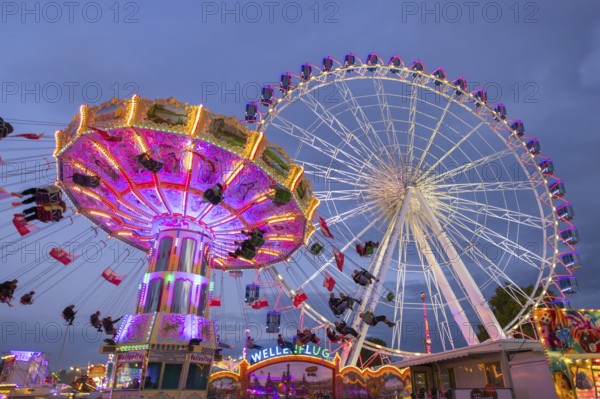A lively funfair at dusk, illuminated by a large Ferris wheel and a colourful chain carousel,