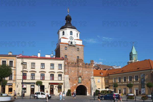 Historic city gate with brick tower and turrets in front of a lively square with passers-by, Krakow