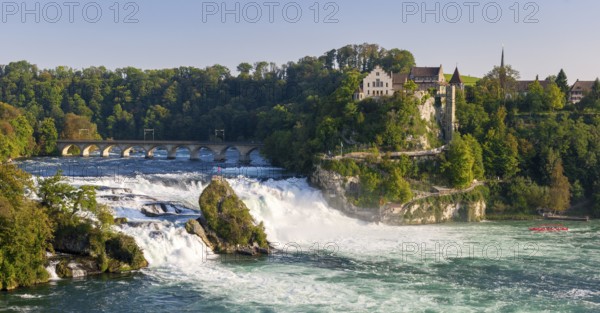 Waterfall flowing next to a historic bridge with lush vegetation and buildings in the background,