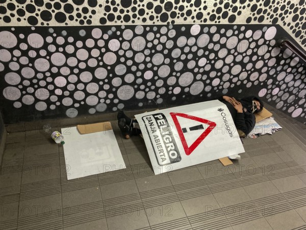 Homeless man sleeping in the entrance of the underground covered with a warning sign reading