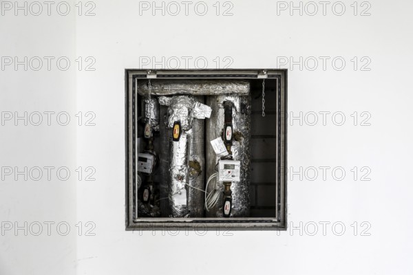 Insulated pipes and measuring device devices of a heating system can be seen behind a white wall,