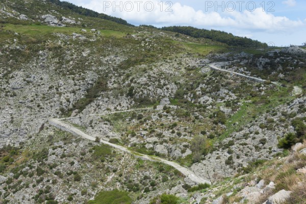 A winding serpentine road leads through a rocky mountain landscape with green grassy areas under a