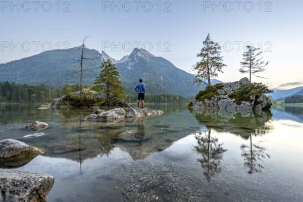 Young man standing on stones in the lake, Hochkalter reflected in Hintersee, at sunset,