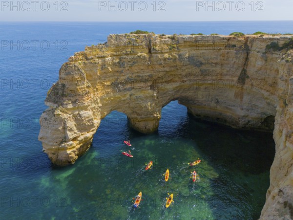 A rocky cliff juts out into the turquoise sea, where several colourful kayaks paddle on the water,