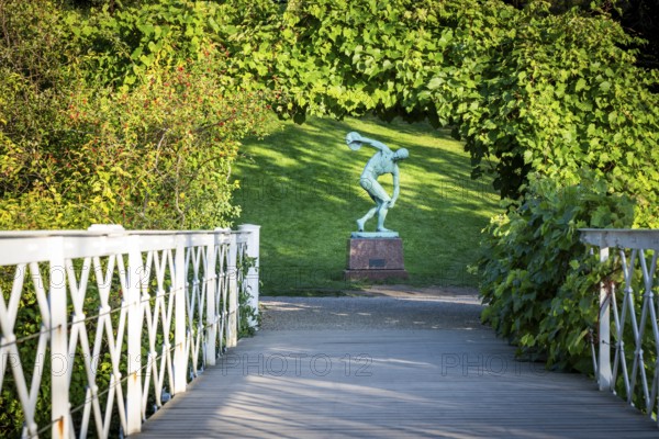 Wooden bridge leading to the sculpture of a discus launcher in a green, sunny park area, Botanical
