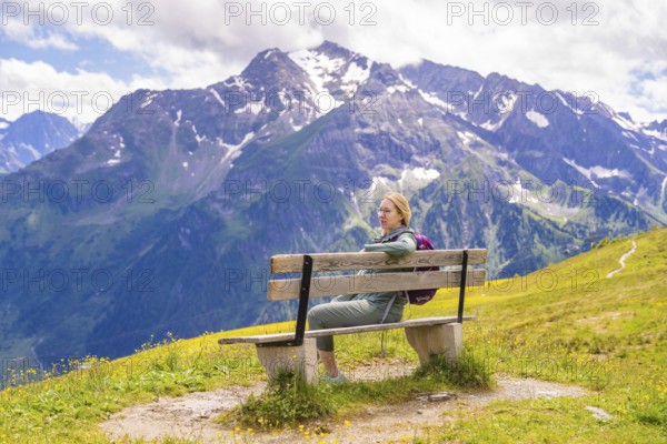 A woman sits on a bench with a view of the snow-covered mountains and the hiking trail, Penken,