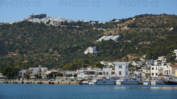 Coastal town with harbour, surrounded by white buildings, at the base of green hills and blue sky,