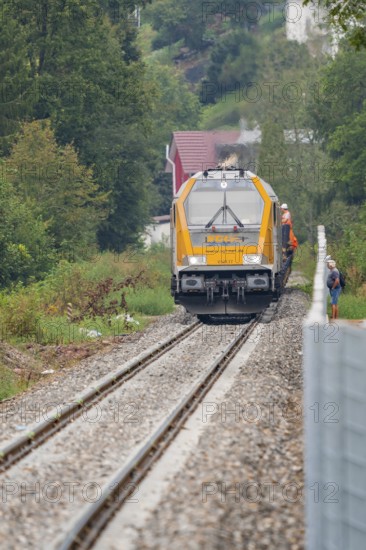 A yellow train runs on rails through a wooded landscape, surrounded by trees and accompanied by