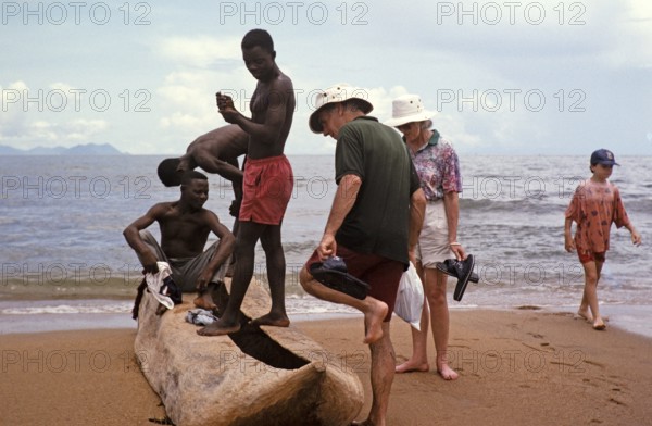 Tourists with local young men looking at a dugout tree on the beach of Lake Malawi, Malawi,