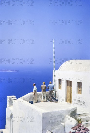 Three male men tourists look out to sea from terrace of whitewashed building at Oia, Santorini,