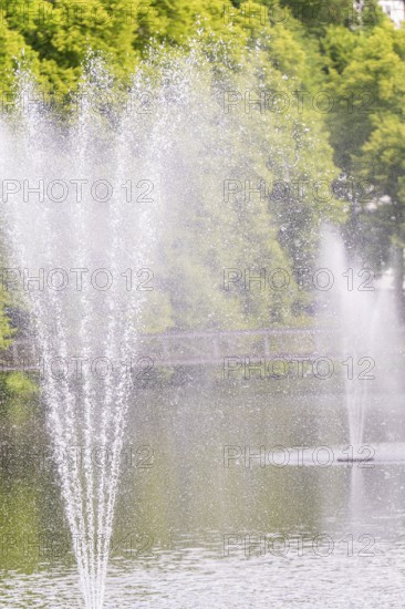 A water fountain sprays high into the air over a calm lake with a green background, Stadtpark Kleb,