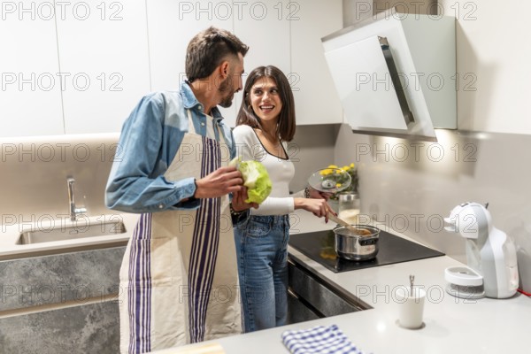 Happy couple wearing aprons, preparing healthy meal together in their modern kitchen, enjoying